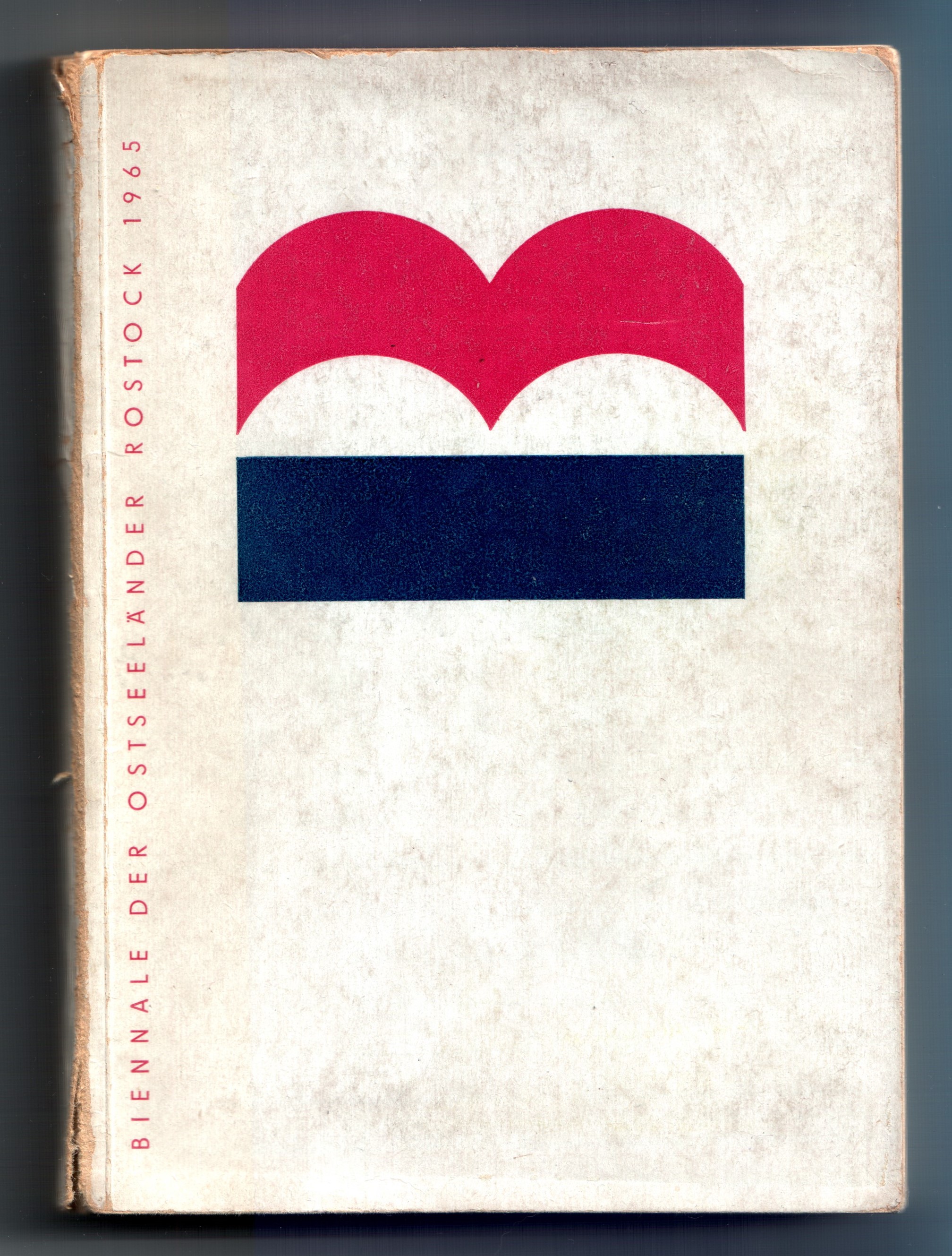 Zu sehen ist ein Foto des Katalogs mit dem Titel „Biennale der Ostseeländer Rostock 1965“ in roter Schrift auf weißem Grund. Auf der oberen Hälfte ist ein großes stilisiertes „B“ abgebildet, das auf dem Rücken liegt und aus einem dunkelblauen Balken unter zwei roten Bögen besteht. Der Umschlag zeigt starke Gebrauchsspuren.